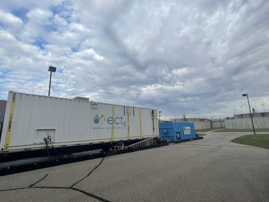 A large white shipping container labeled ect2 Montana Environmental Group and a blue unit provide PFAS solutions, set up on a paved area beside a fenced facility under a cloudy sky.