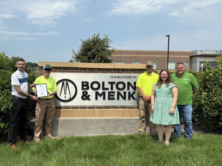 Five people stand in front of a Bolton & Menk sign outdoors; two hold a Military Outreach plaque or certificate. Three wear yellow or green shirts, one wears a dress, and one holds the certificate. A brick building and greenery are in the background.