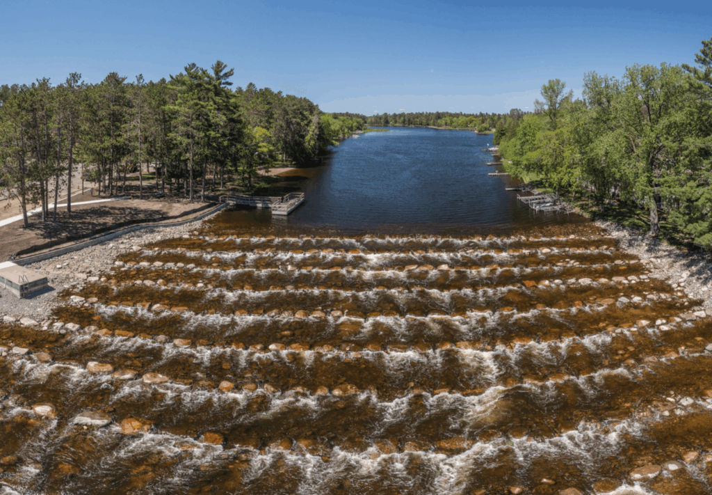 A river flows over several rows of rocks, creating small rapids, surrounded by dense green trees under a clear blue sky. Upstream near the docks along the forested shoreline, Pine River Dam Renewal work enhances the area's natural beauty.