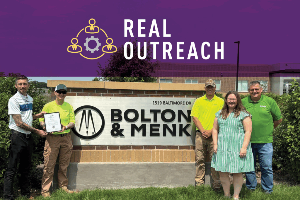 Five people stand smiling in front of a “Bolton & Menk” sign. One person hands an award to another. Above them is a purple banner with a yellow icon and white text reading “REAL OUTREACH.”.