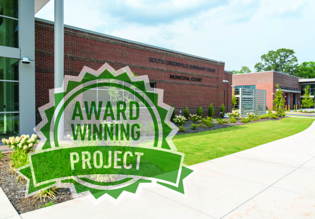 A modern brick municipal building with landscaped grounds and a large graphic in the foreground that reads AWARD WINNING PROJECT in green letters inside a badge-shaped design.