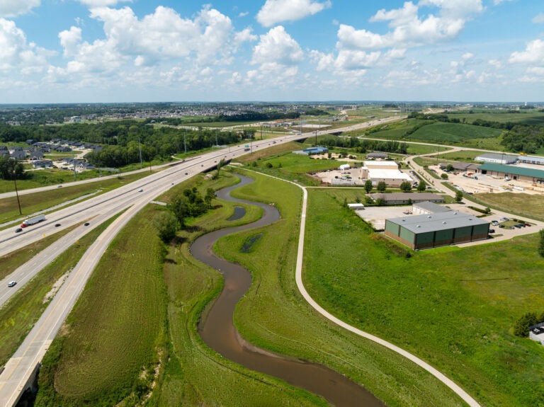 Aerial view of a highway curving through a green landscape with a winding creek, scattered buildings, and open fields under a partly cloudy sky near a stream mitigation bank.