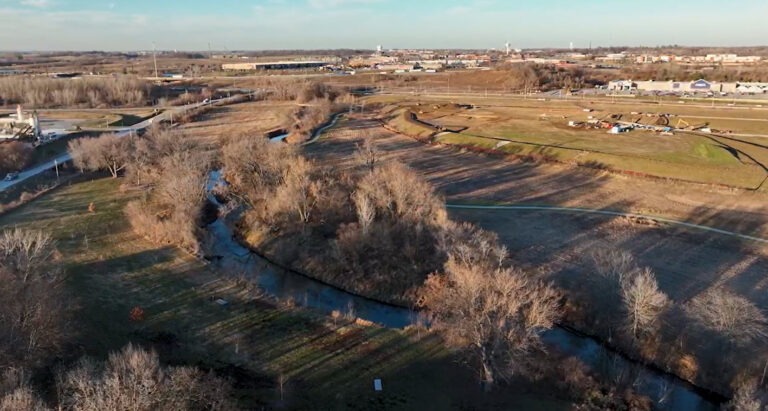 Aerial view of a winding creek, part of a stream mitigation bank, bordered by leafless trees running through open fields, with a town and buildings visible in the background under a clear sky.