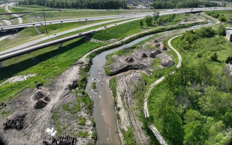 Aerial view of a muddy creek winding through green grassy areas and construction mounds within a stream mitigation bank, with scattered debris, a highway overpass, and cars in the background. Trees and walking paths line the scene’s edges.