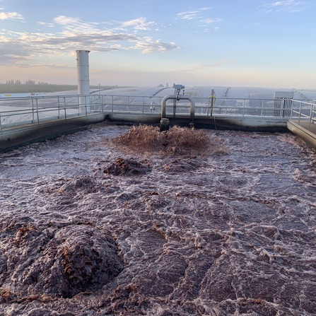 Aeration tank with frothy, reddish-brown wastewater and bubbling surface at a treatment facility under a clear sky.