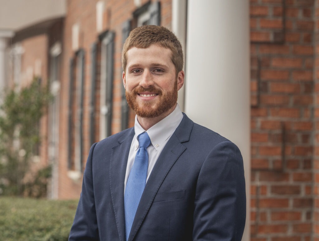 A young man with short brown hair and a beard, wearing a navy blue suit, white shirt, and blue tie, stands smiling in front of a brick building with white columns and greenery.