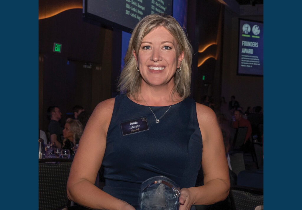 A woman with blonde hair in a navy dress smiles and holds an award at an indoor event.