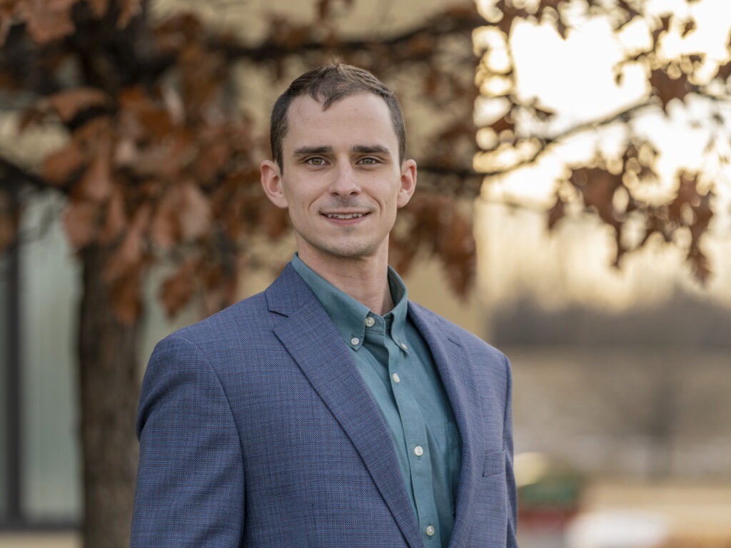 A man wearing a blue blazer and green shirt stands outdoors in front of a tree with brown leaves, smiling at the camera. The softly blurred background hints at natural light, evoking the thoughtful planning seen in Snow Storage Design.