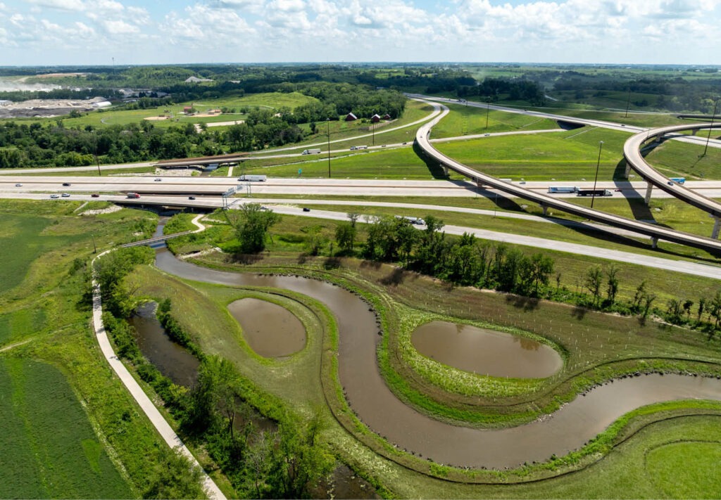 Aerial view of a highway interchange with cars, curving roads, green grassy areas, and wetlands with ponds below, surrounded by trees and pathways under a partly cloudy sky.