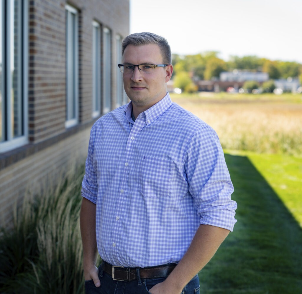 A man wearing glasses and a light purple checkered shirt stands outside beside a brick building with his hands in his pockets, looking at the camera. Behind him is grass, a field, and land used for stream mitigation bank purposes.