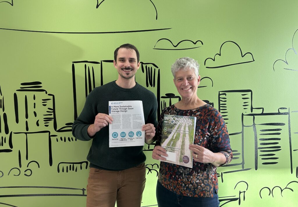 Two people stand smiling in front of a green wall with a cityscape illustration. The man holds a printed document, and the woman holds a magazine titled ENVIRONMENTAL CONNECTION.