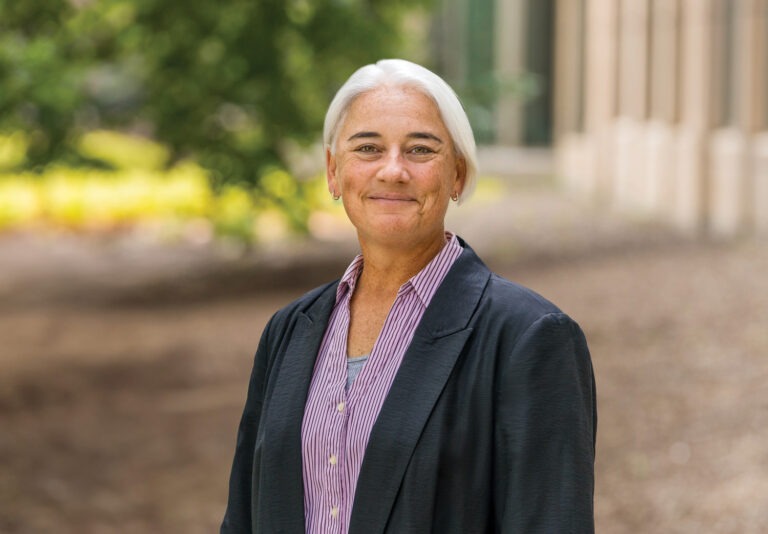 An older person with short white hair, on the Board, wears a black blazer over a striped shirt and stands outdoors smiling at the camera, with greenery and a building visible in the blurred background.