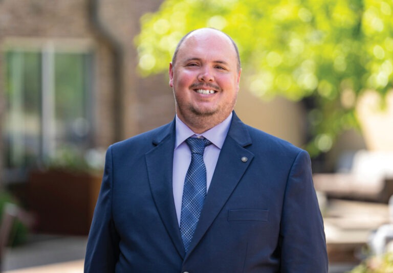 A man in a navy suit and tie, smiles outdoors. He is bald and stands before blurred greenery and buildings, with sunlight creating a bright, pleasant background.