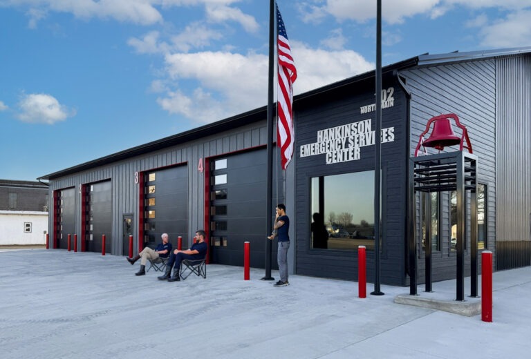 Three people sit and stand outside a modern black building labeled “Hankinson Emergency Services Center.” An American flag and a red fire bell tower are in front. The area is paved, with blue sky and clouds overhead.