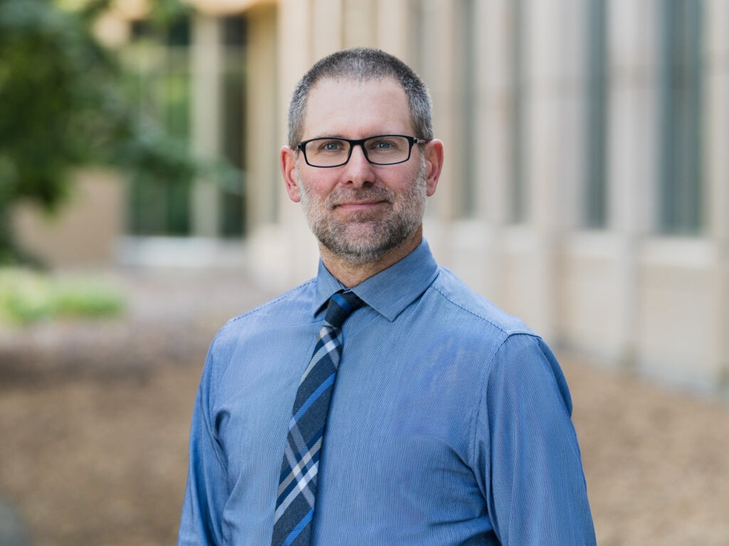 A man with short hair, a beard, and glasses wearing a blue shirt and plaid tie stands outdoors.