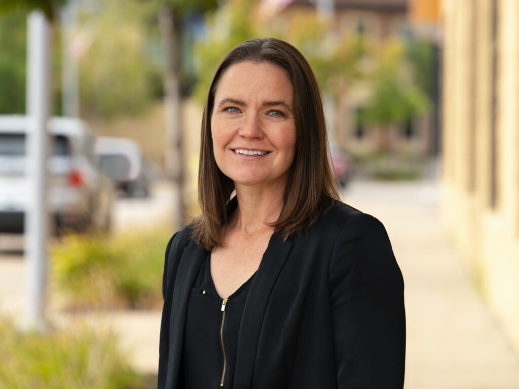 A woman with straight brown hair wearing a black blazer and top smiles while standing outdoors on a city sidewalk, with greenery, building in the background.