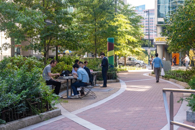 People sit and talk at outdoor tables along a winding brick path surrounded by greenery in an urban park. Several people stroll through Bearden Promenade, while tall buildings and cars are visible in the background.