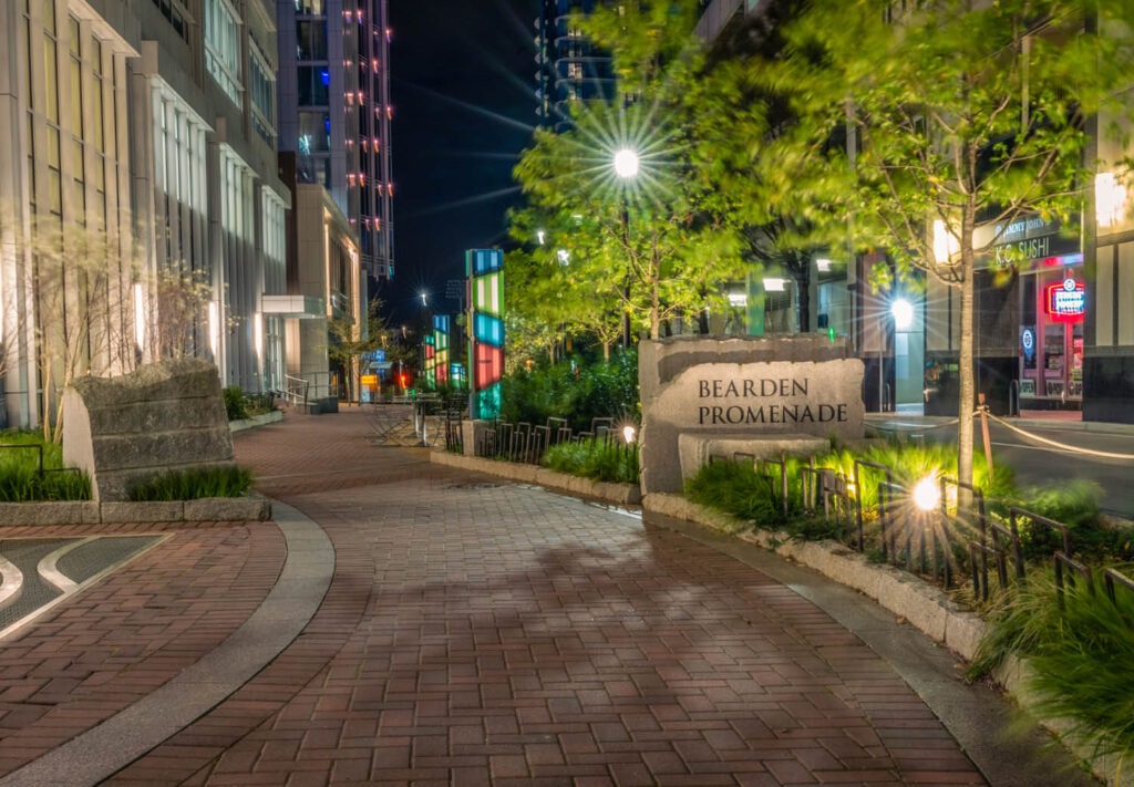 Night view of Bearden Promenade with curved brick pathway, illuminated buildings, green trees, a lit sign, and colorful lights on the left, creating a vibrant urban atmosphere.