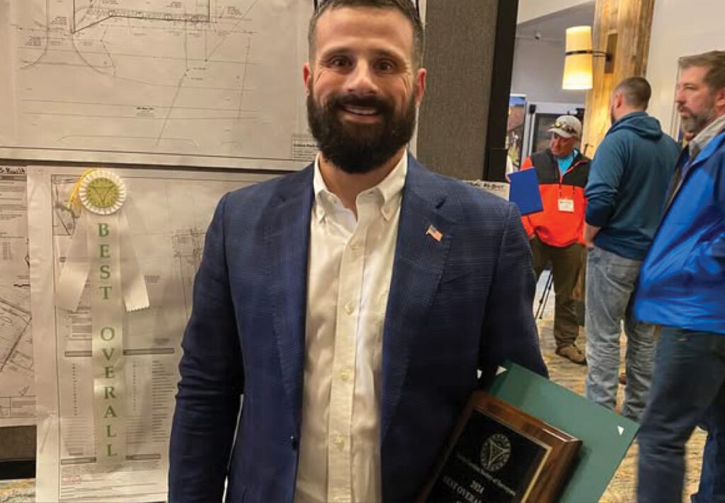 A man in a navy blazer holds a plaque and stands in front of a map with a Best Overall ribbon. Several people are in the background in a room with wooden decor.