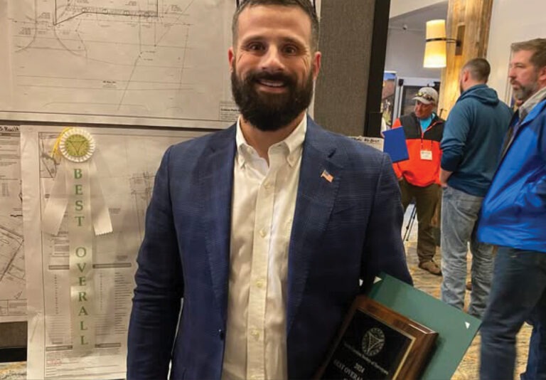 A man in a navy blazer holds a plaque and stands in front of a map with a Best Overall ribbon. Several people are in the background in a room with wooden decor.