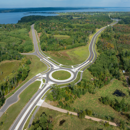Aerial view of a roundabout surrounded by forested landscape, open fields, and a nearby lake, with multiple roadways connecting through the intersection.