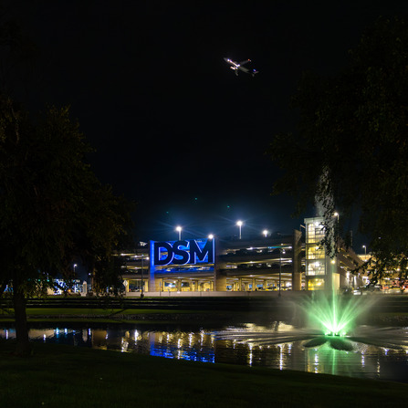 Des Moines airport parking structure at night with illuminated DSM sign, green-lit fountain, and airplane overhead.