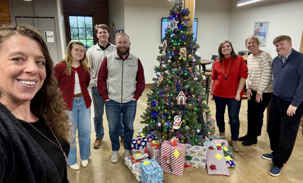 Seven people stand smiling around a decorated Christmas tree with wrapped presents beneath it, capturing the joy of a holiday gathering.
