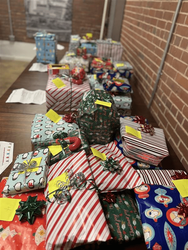 A table covered with brightly wrapped holiday presents, showcasing festive designs and bows, captures the Season of Giving Back. Against a brick wall, papers and sticky notes adorn some gifts, adding extra thoughtfulness to the holiday spirit.