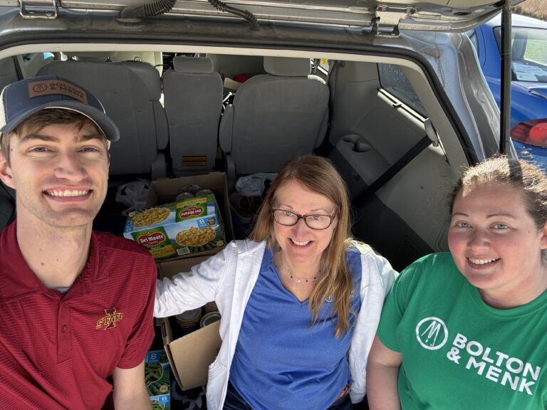 Three people smile at the camera while sitting at the open back of a vehicle filled with boxes of food, embodying the Season of Giving Back as they participate in a food drive or donation event.