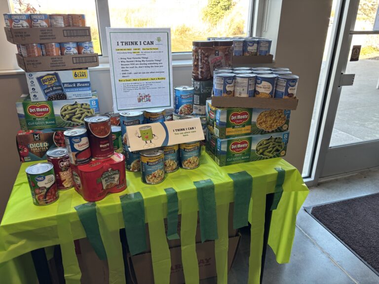 A table covered with a green tablecloth holds stacks of canned vegetables and beans. A sign reads I THINK I CAN, featuring a small cartoon train.