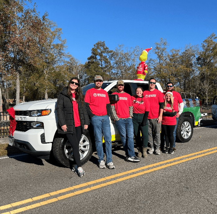 Seven people in matching red shirts stand smiling in front of a decorated white pickup truck on a sunny day. A holiday figure sits on the truck bed, with trees lining the background.