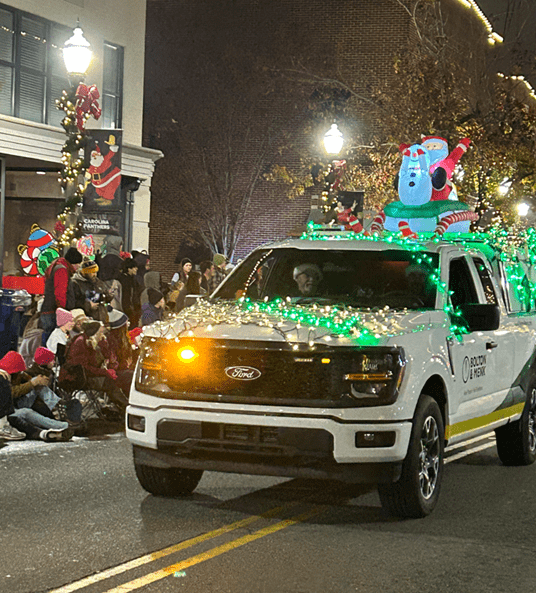 A white pickup truck decorated with green Christmas lights and inflatable holiday figures drives in a nighttime parade as people in festive attire watch from the sidewalk.