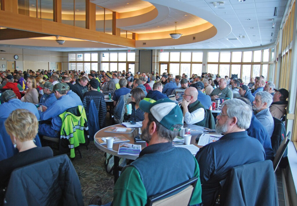 A large group of people, mostly men, sit at round tables in a bright conference room. Many wear casual or work attire, and some have hats. The attentive atmosphere suggests a Salt Symposium meeting or seminar in progress.