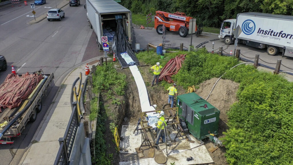 Aerial view of construction workers in safety vests repairing an underground pipe next to a busy road, with trucks, equipment, and materials surrounding the excavation site.