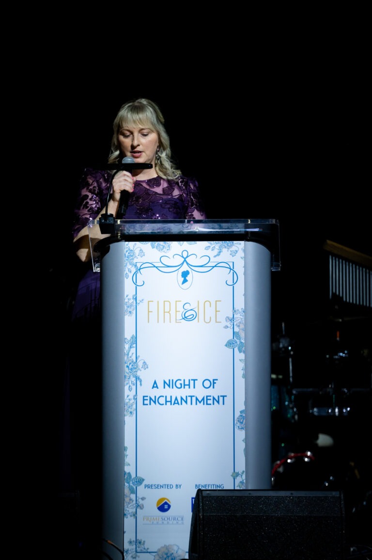 A woman stands at a podium decorated with floral designs and the text Fire & Ice: A Night of Enchantment, speaking into a microphone at this Season of Giving Back event. The background is dark, and she is illuminated by stage lighting.