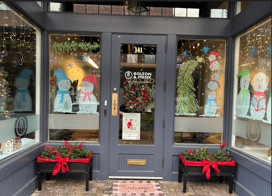 A storefront decorated for the holidays with snowman paintings on the windows, a wreath on the door, hanging lights, and planters filled with greenery and red bows. A welcome mat sits at the entrance.