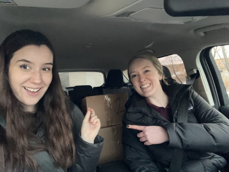 Two smiling women sit in the front seats of a car, pointing at cardboard boxes stacked in the back seat.