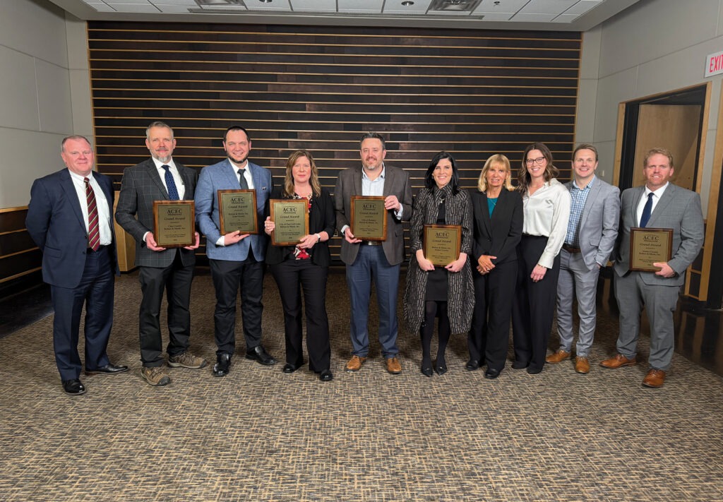A group of ten professionally dressed people stand indoors, smiling, with six holding plaques or awards. The background features wood paneling and carpeting typical of a conference or banquet room.