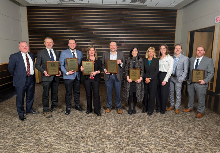 A group of ten professionally dressed people stand indoors, smiling, with six holding plaques or awards. The background features wood paneling and carpeting typical of a conference or banquet room.