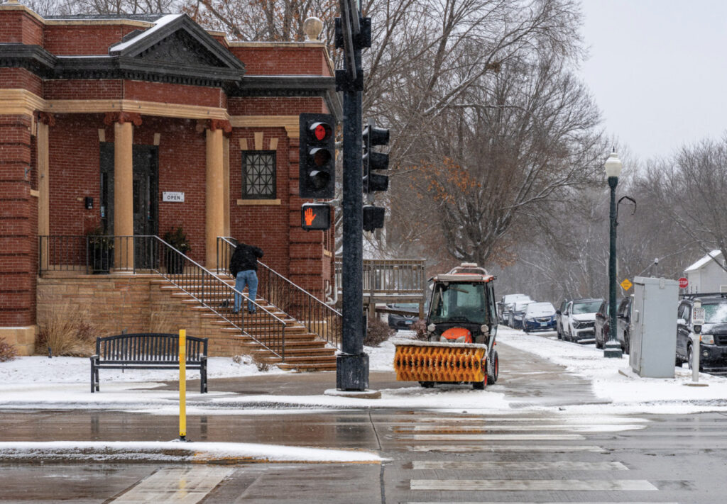A person shovels steps outside a brick building on a snowy day, while a small snowplow is parked on the sidewalk near a traffic light. Snow covers the ground and nearby parked cars.