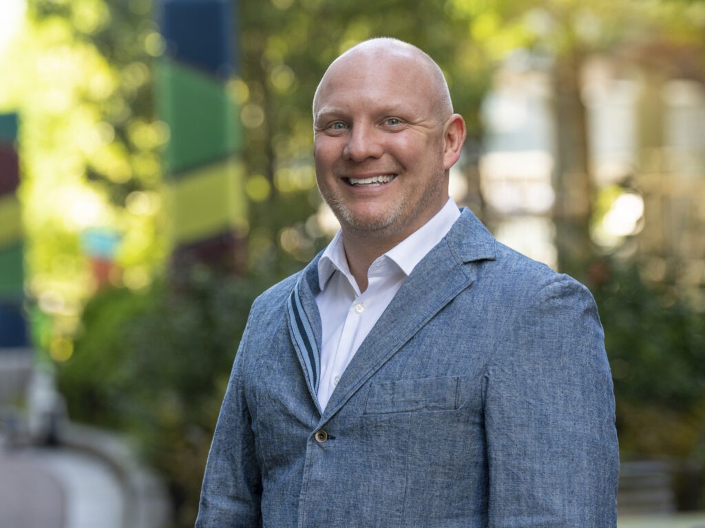 A smiling bald man in a blue blazer and white shirt stands outdoors, with greenery and colorful poles in the blurred background.