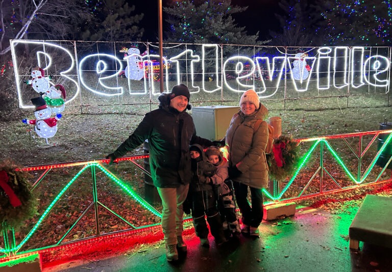 A family of four in winter clothes stands smiling in front of a large illuminated Bentleyville sign decorated with snowmen and colorful lights at night.