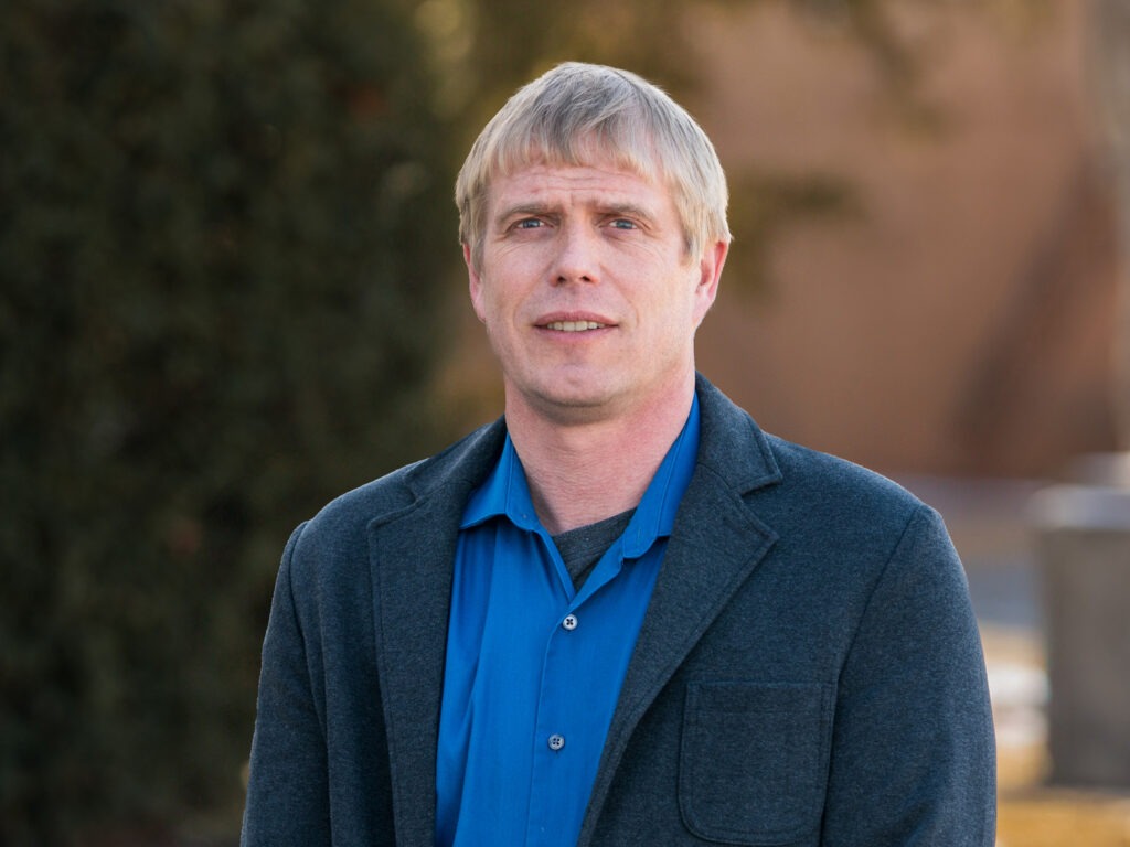 A man with short blond hair wearing a blue shirt and dark blazer stands outdoors, looking at the camera.