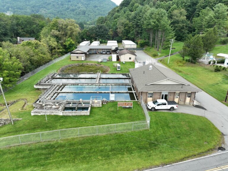 Aerial view of a water treatment plant, showcasing vital water infrastructure funding with rectangular tanks, a fenced grassy area, an office building, a white truck parked outside, and surrounding trees and hills in the background.