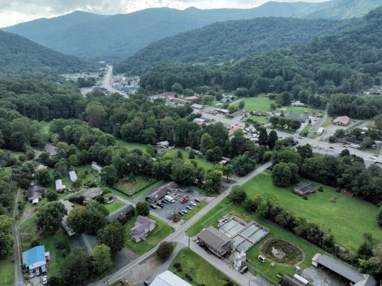Aerial view of a small town surrounded by lush green hills and forests, with scattered houses, buildings, and evidence of water infrastructure funding supporting modern roads and parking lots winding through the landscape under a cloudy sky.