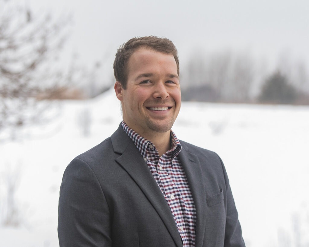 A man in a checked shirt and dark blazer, smiles while standing outdoors in a snowy landscape with bare trees in the background.