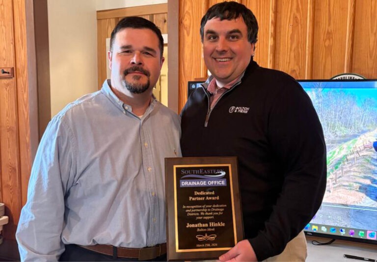 Two men standing indoors, one holding a plaque that reads Dedicated Partner Award. Both are smiling at the camera. Wood-paneled walls and a television displaying an outdoor scene are in the background.