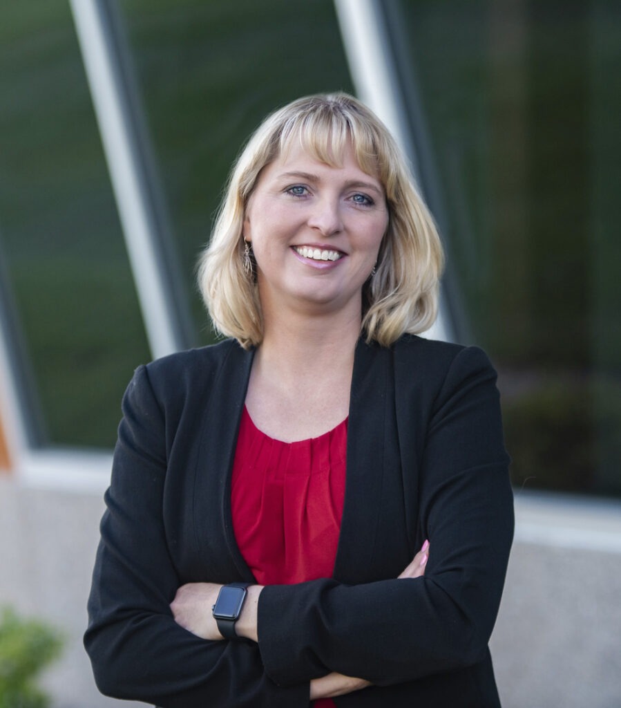A woman with blonde hair, wearing a black blazer and red top, stands outdoors with arms crossed, smiling confidently at the camera.