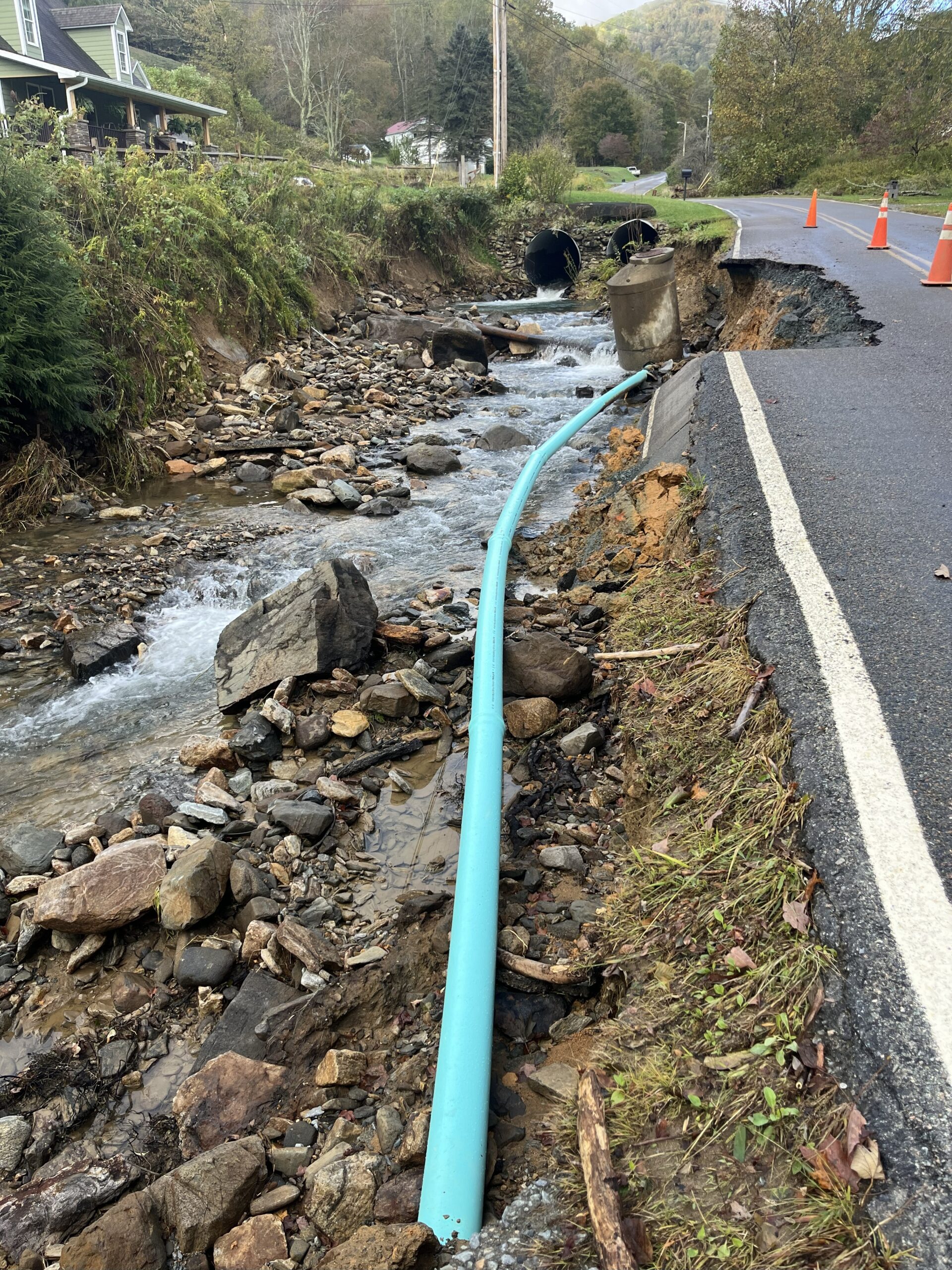 A damaged roadside shows erosion from a stream, exposing a blue pipe and crumbling pavement—a stark reminder of the need for water infrastructure funding. Traffic cones mark the washed-out area near a rural house and trees.