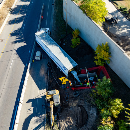 Aerial view of a deep, shored excavation next to an active freeway, with safety fencing and temporary traffic control in place.